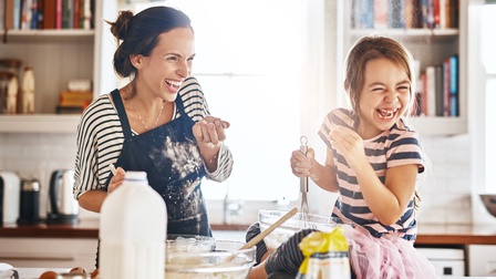 Eine erwachsene Person kocht spielerisch mit einem Kind in einer Küche und beide lachen