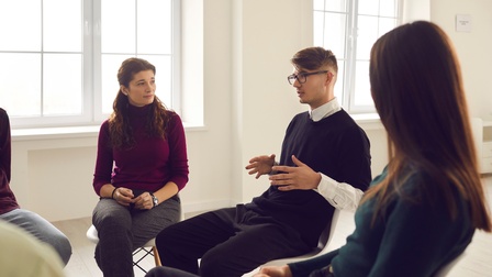 In einem Sesselkreis sitzen mehrere Personen. Eine der Personen mit Brille spricht. Die anderen blicken zu ihr