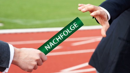 In der rechten Bildhälfte ist ein sich ausstreckender Arm in einem Sakko einer Person. In der linken Bildhälfte ist ein Arm in einem Sakko, die einen Stab in Richtung der anderen Hand hält. Auf dem Stab steht der Schriftzug Nachfolge