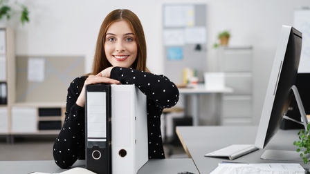 Eine Person steht an einem grauen Schreibtisch in einem hell und steril eingerichteten Büro. Ihre Arme sind an zwei Ordner gelehnt, vor ihr liegt ein Notizbuch und ein Kugelschreiber, neben ihr ein Monitor, Dokumente und eine Fernbedienung. 