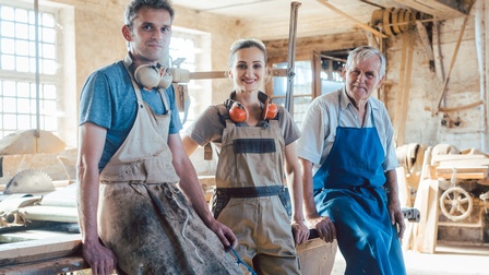 Drei sanft lächelnde Personen unterschiedlicher Generationen in Arbeitskleidung stehen in Tischlerei