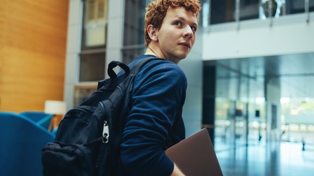 Eine Person mit einem Laptop und Rucksack betritt ein Universitätsgebäude und blickt nach hinten.