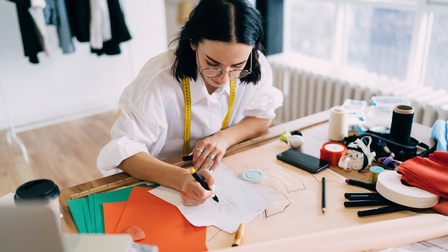 Person mit kurzen dunklen Haaren und Brille sowie weißer Bluse mit umgehängten Maßband sitzt an einem Tisch und zeichnet ein Schnittmuster, am Tisch liegen weitere Nähutensilien, im Hintergrund steht ein voller Kleiderständer