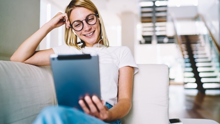 Lächelnde Person mit Brillen und Kopfhörern  sitzt auf Couch, stützt einen Arm an den Kopf, hält in anderer Hand ein Tablet