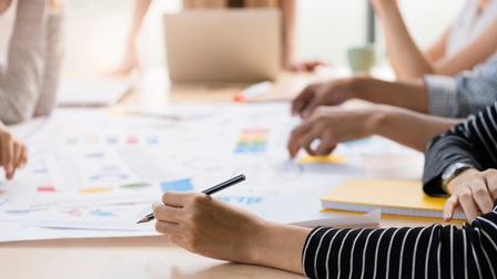 Um einen Tisch sitzen mehrere Menschen. Eine Person steht am Tisch. Auf dem Tisch liegt ein großes, weißes Plakat mit verschiedenen Begriffen und farblich gekennzeichneten Bereichen