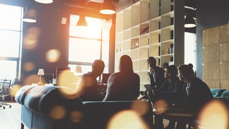Gruppe von Personen sitzt rund um einen Tisch in einem Büro und hält eine Besprechung. Die Sonne scheint so durch das Fenster, dass nur die Silhouetten der Personen zu erkennen sind.