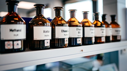 Braune Glasflaschen mit weißen Etiketten und schwarzen Aufschriften nebeneinander in Regal stehend, im Fokus Apothekerflasche mit Schriftzug Ethanol und Symbol einer Flamme