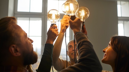 Personen stehen in einer Gruppe zusammen und halten freudig leuchtende  Vintage-Glühbirnen mit Kabel in die Höhe des Raumes