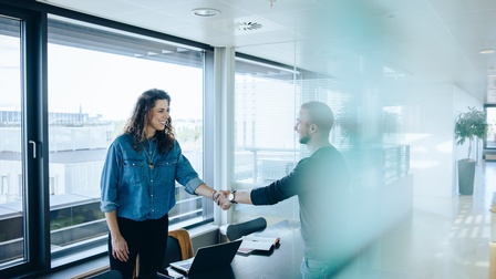 Zwei lächelnde Personen reichen sich in Besprechungsraum über Tisch hinweg die Hand