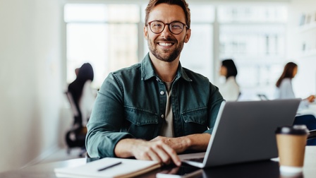 Nahaufnahme einer Person mit Brille, die vor einem aufgeklappten Laptop sitzt, der auf einem braunen Bürotisch steht. Am Tisch ist außerdem ein Coffee-to-go-Behälter, ein Smartphone und ein Notizblock mit einem Stift 