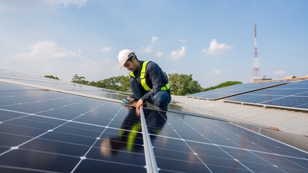 Auf einem leicht schrägen Dach sind viele dunkle Platten montiert. Die Platten werden von einem Metallrahmen umfasst. Auf den Platten ist eine Person mit Helm. Sie hält einen Bohrer in der Hand. Der Himmel ist blau und leicht bewölkt