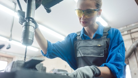 Eine Person mit braunen zurückgebundenen Haaren, einer Schutzbrille und Arbeitshandschuhen, verwendet eine Ständerbohrmaschine und beugt sich leicht über ihre Arbeit