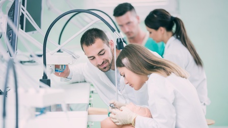 Personen stehen rund um eine Maschine, mit der Zähne modelliert werden können, eine bedient diese. Die Personen sind in einem Labor, in dem technische Geräte zu sehen sind. 