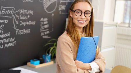 Person mit langen braunen Haaren und Brille hält eine Mappe und steht freudig vor einer Tafel mit Notizen