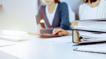 Mappen liegen gestapelt auf einem Tisch, im Hintergrund sitzen zwei Personen vor einem Monitor in der Unschärfe