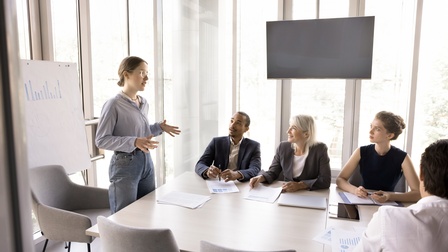 Seitliche Aufnahme einer an einem Tisch stehenden Person. An dem Tisch sitzen vier Personen, vor denen unterschiedlich bedruckte Zettel liegen. Die sitzenden Personen blicken zur stehenden Person. Hinter dieser ist eine Tafel mit einer Statistik