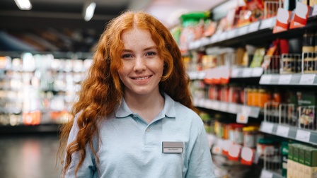 Porträt einer sanft lächelnden Person mit langen, rötlichen Haaren in einem hellen Oberteil und Namensschild auf dem Nina Petersen steht. Rechts neben der Person ist ein mit Lebensmitteln befülltes Regal.