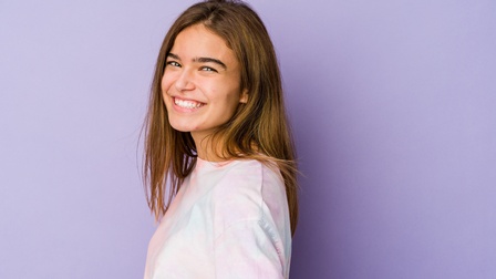 Seitliches Porträt einer Person mit offenen, braunen Haaren vor einer lila Wand. Die Person trägt ein helles Oberteil und hat den Mund leicht geöffnet