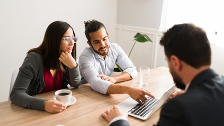 An einem Tisch sitzt eine Person in einem Sakko. Der Person gegenüber sitzen zwei Personen. Zwischen ihnen steht ein aufgeklappter Laptop, auf den eine Person deutet. Die andere Person hat eine Tasse Kaffee vor sich