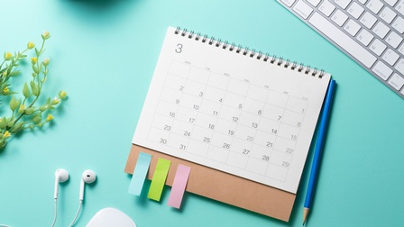 Kalender mit Monatsblatt liegt auf einem türkisen Untergrund, eine Tastatur sowie eine Pflanze, Kopfhörer, Maus und ein Bleistift liegen daneben, Topshot