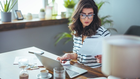 Person mit dunklen langen Haaren und Brille sitzt an einem Tisch, blickt auf Unterlagen in der Hand und hält mit der anderen Hand einen Stift, vor ihr steht ein aufgeklappter Laptop sowie eine Tasse, ein Glas, eine Sanduhr und Unterlagen in einem Wohnraum