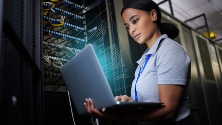 Person blickt in aufgeklapptes Notebook aus dem Hologramm von Computercodes empor steigen, im Hintergrund Serverracks