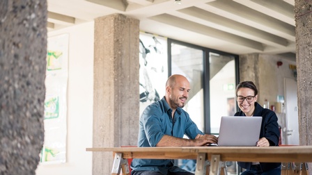zwei Personen sitzen an einem Holztisch und schauen auf den Bildschirm eines Laptops