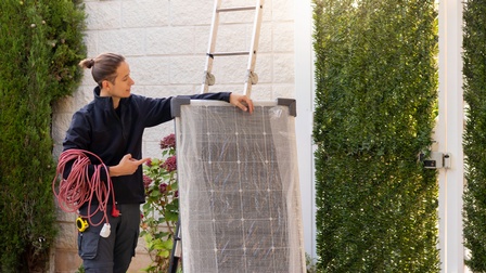 Eine Person in Arbeitskleidung hat ein langes Kabel um den rechten Arm gewickelt. Mit dem linken Arm hält sie eine verpackte Solarzelle. Neben der Solarzelle ist die Wand eines Gebäudes, an das eine Leiter angelehnt ist