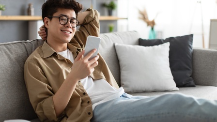 Jugendliche Person mit dunklen kurzen Haaren und Brille, sowie beigem Hemd, weißem Shirt und Jeans sitzt freudig auf einer Couch und blickt auf ein Smartphone