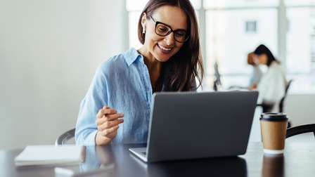Nahaufnahme einer Person mit Brille, die vor einem aufgeklappten Laptop sitzt. Der Laptop steht auf einem dunkelbraunen Holztisch, daneben steht ein Coffee-to-go-Becher, ebenso liegen ein Notizblock und ein Smartphone auf dem Tisch
