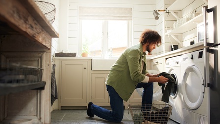 Bärtige Person mit Locken kniet mit einem Bein am Boden, das andere angewinkelt aufgestellt, vor Waschmaschine und hält Wäsche in Händen, daneben Wäschetrockner, vor Person Drahtkorb mit Wäsche, ringsum Waschküchenregale und Fenster