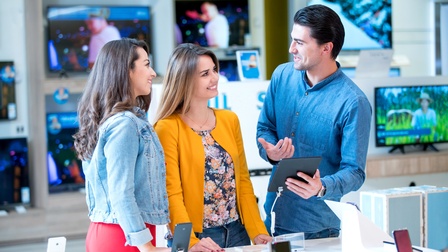 Drei lächelnde Personen in Gespräch, eine Person in erklärender Haltung mit Tablet in Hand, ringsum Smartphones und Fernseher auf Verkaufsregalen