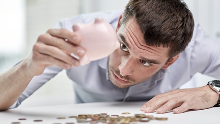 Person mit kurzen dunklen Haaren blickt erwartungsvoll auf ein ausgeleertes  Sparschwein während Münzen auf dem Tisch liegen