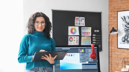 Porträt einer Person mit lockigen, dunklen Haare und Brille, die ein Tablet hält. Hinter der Person sind ein Bildschirm und ein aufgeklappter Laptop. Auf dem Bildschirm ist ein Programm geöffnet zur Bearbeitung von Dateien