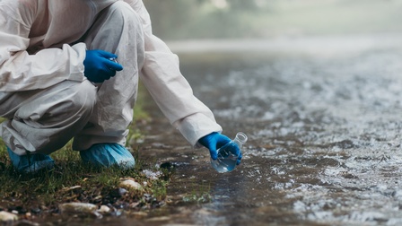 Eine Person in Schutzkleidung kniet neben einem Geewässer und entnimmt mit einer kleinen Flasche eine Wasserprobe. Die Person wird links vom Bildrand übschnitten, rechts ist das Gewässer zu sehen.