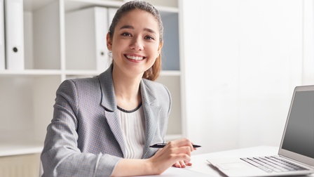 Porträt einer an einem Tisch sitzenden Tisch. Vor ihr liegt ein Clipboard. Sie hält einen Kugelschreiber in der rechten Hand. Schräg links vor ihr steht ein aufgeklappter Laptop. Hinter ihr ist ein weißes Regal