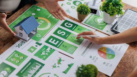 Auf einem Tisch liegen mehrere Zettel mit Diagrammen und Abbildungen. Auf mehreren Zettel stehen die Buchstaben ESG. Am Tisch stehen sich zwei Personen gegenüber. Die rechte Person deutet mit einem Stift auf den Buchstaben S