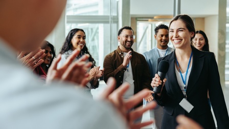 Personen mit Ausweisschildern stehen in einem Halbkreis und klatschen. Im Kreis steht eine Person mit dunklem Sakko, Mikrofon sowie Ausweisschild, die lächelt