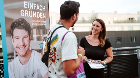 Zwei Personen, die sich anlächeln, dahinter ein Rollup mit dem Schriftzug 