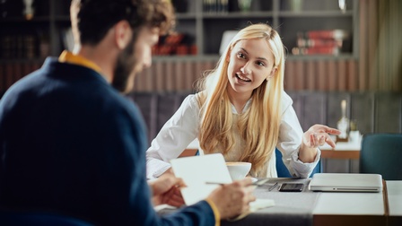 Rückenansicht einer Person, die in einem Lokal an einem Tisch sitzt und etwas in einem Notizblock notiert. Ihr gegenüber sitzt eine weitere Person. Die beiden sitzen sich gegenüber und reden miteinander