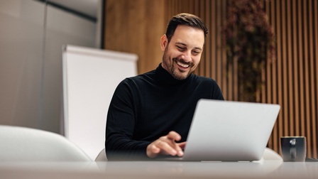 Person mit kurzen Haaren und Bart sitzt bei einem Tisch und arbeitet freudig mit einem Laptop, im Hintergrund zeigt sich eine Holzvertäfelung sowie ein Flipchart