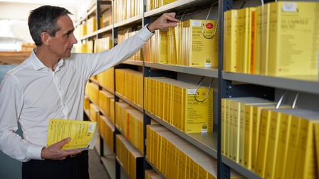 Person mit weißem Hemd hält ein Rechtsbuch in Händen und greift mit der anderen Hand zu einem weiteren Buch in einem Regal einer Bibliothek