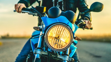 Nahaufnahme des vorderen Teils und des Scheinwerfers eines Motorrads. Auf dem Motorrad sitzt eine Person. Im Hintergrund ist eine asphaltierte Straße.