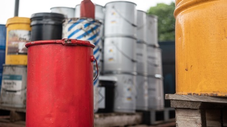 Roter Metalleimer im Fokus, nebenstehend im Ausschnitt gelbes Metallfass auf Holzpalette im Hintergrund verschwommen weitere Fässer neben- und aufeinander gestapelt