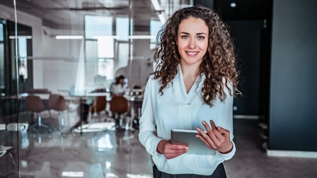 Person mit lockigem langen Haar, blauer Bluse blickt freudig in die Kamera, während sie ein Tablet in ihren Händen hält, im Hintergrund zeigt sich ein modernes, gläsernes Büro