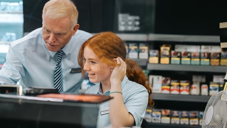 Eine Person in einem hellblauen Oberteil und mit Namensschild, auf dem Nina Petersen steht, sitzt. Links neben ihr ist eine weitere Person. Beide blicken leicht nach unten. Hinter ihnen ist ein mit Objekten befülltes Regal.