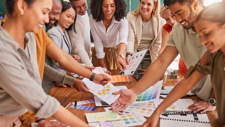 Mehrere Personen stehen freudig rund um einen Besprechungstisch, halten und zeigen auf Farbkarten und weitere Unterlagen