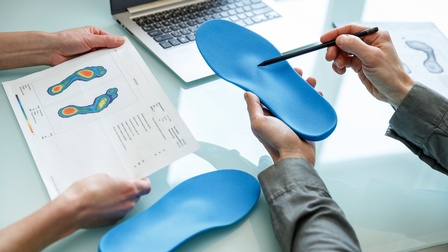 Detailansicht zweier Handpaare: Eines hält Blatt mit Vermessungsscan von Füßen in Händen, anderes hält blaue Schuheinlage in Hand und deutet mit Stift auf Erhebung auf Einlage, im Hintergrund aufgeklapptes Notebook