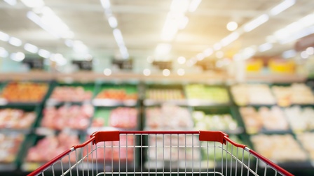 Nahaufnahme des vorderen Teiles eines Einkaufswagens aus Perspektive der fahrenden Person. Im Hintergrund sind verschwommene Kisten, befüllt mit Objekten