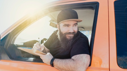 Eine Person mit Kappe, dunklem Oberteil und Bart lehnt sich aus dem Fenster eines orangen Fahrzeugs und notiert mit einem Kugelschreiber etwas auf einem Klemmbrett